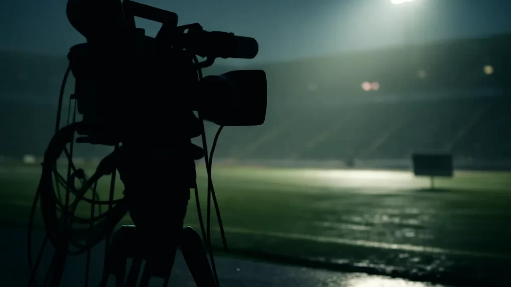 Silhouetted broadcast camera and empty stadium under dusky floodlights.